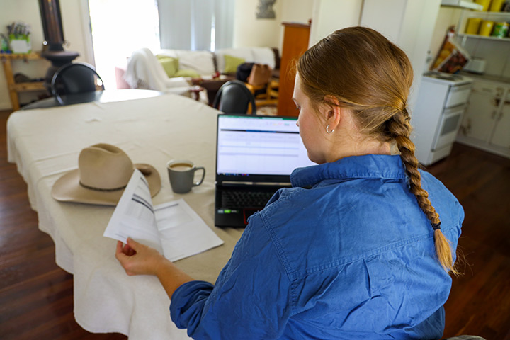 A female farmer wearing a blue workshirt and a single braid sits at a table with a laptop and paperwork in front of her.