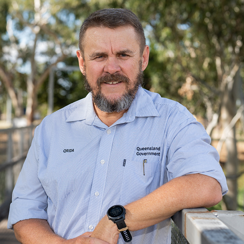 A man with a beard, stands facing the camera smiling. There are gum trees in the background.