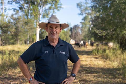 Regional Area Manager John Metelli stands smiling at camera with hands on his hips and cattle grazing in the background. John wears a cream Akubra hat and a navy polo shirt. 